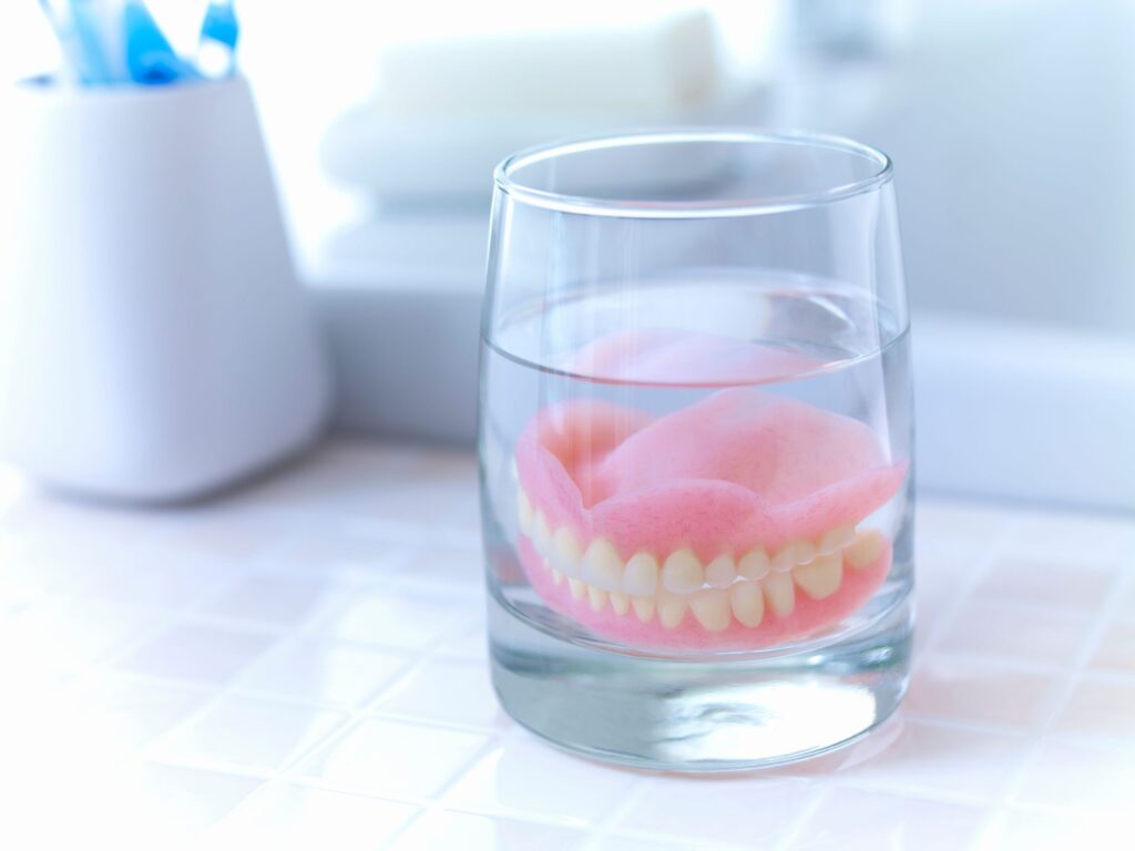 Complete set of dentures in glass of clear fluid on tiled countertop